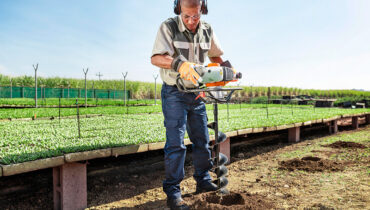 A man using an earth auger to drill holes in the ground for his greenhouse poles.