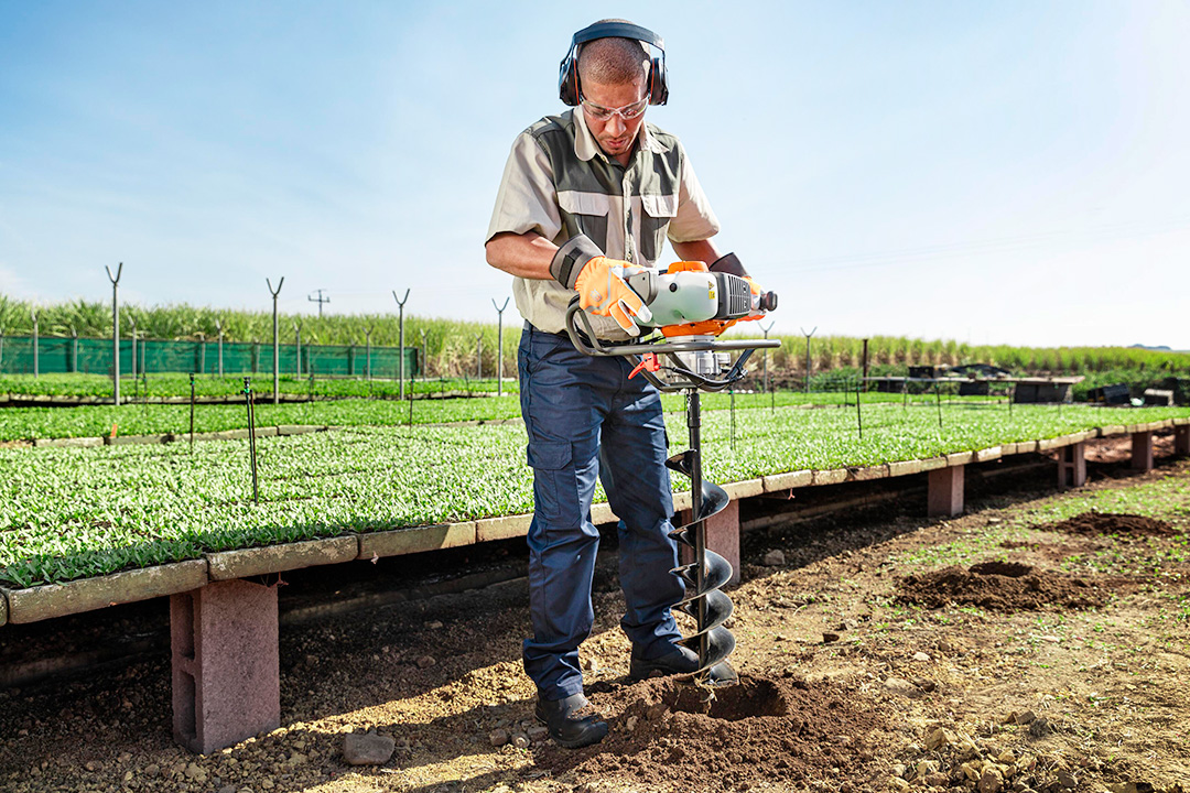 A man using an earth auger to drill holes in the ground for his greenhouse poles.