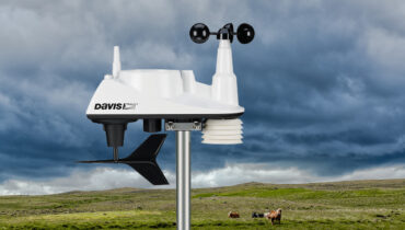 A Davis weather station with a moody sky and farm in the background